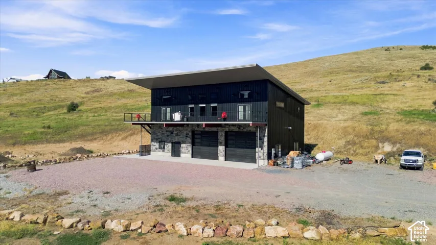 Contemporary exterior with dark siding, stone accents, two garage doors, an upper-level balcony, and a gravel driveway