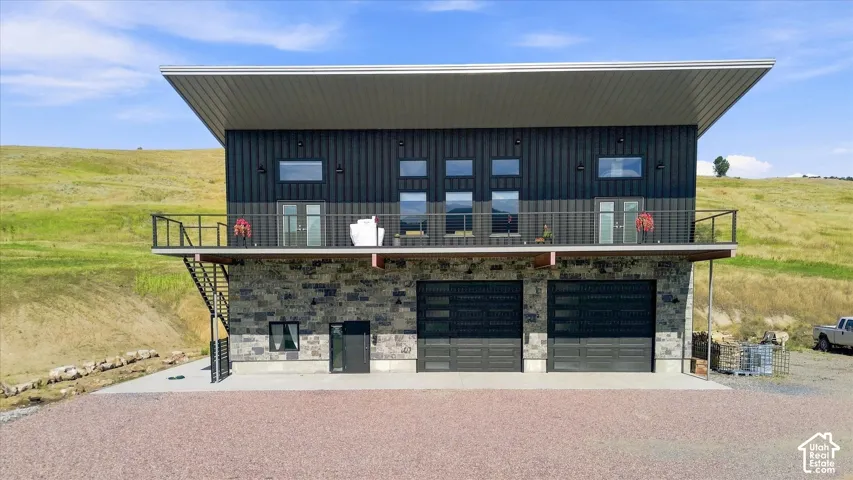 Modern exterior featuring a prominent deck with metal railing, two large garage doors with horizontal window inserts, and a stone facade