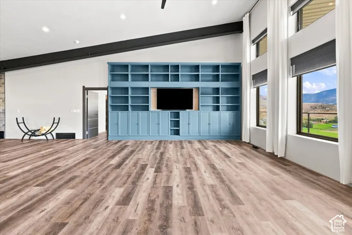 Vaulted ceiling with exposed beams, wood-finish flooring, custom built-in shelving, large windows, and a stone-finish fireplace