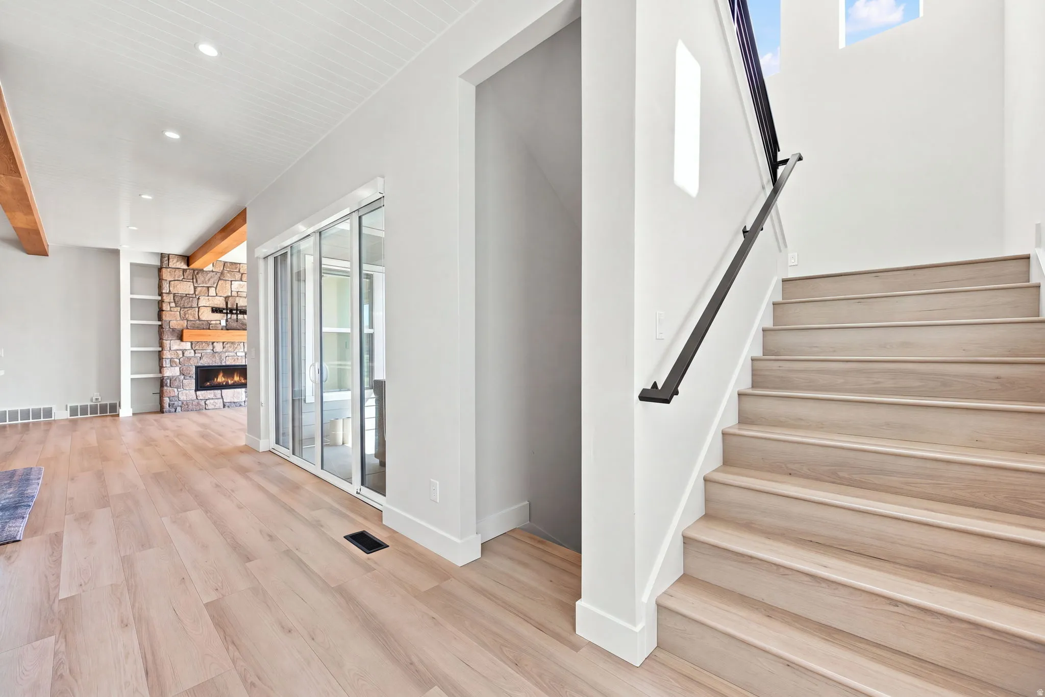 Light wood-finish flooring extending into the stone-faced fireplace area, featuring built-in shelving and exposed wood beams