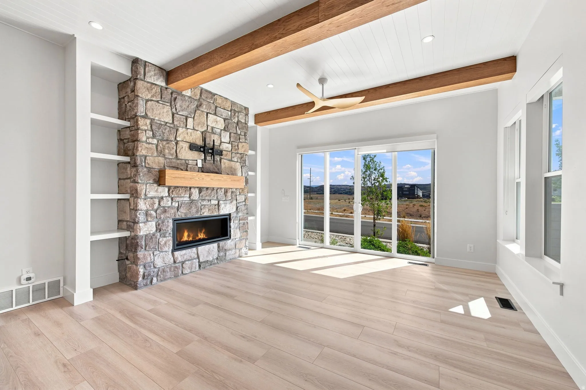 Stone-clad fireplace with built-in shelving, wood-finish flooring, exposed wood beams, and a large sliding glass door