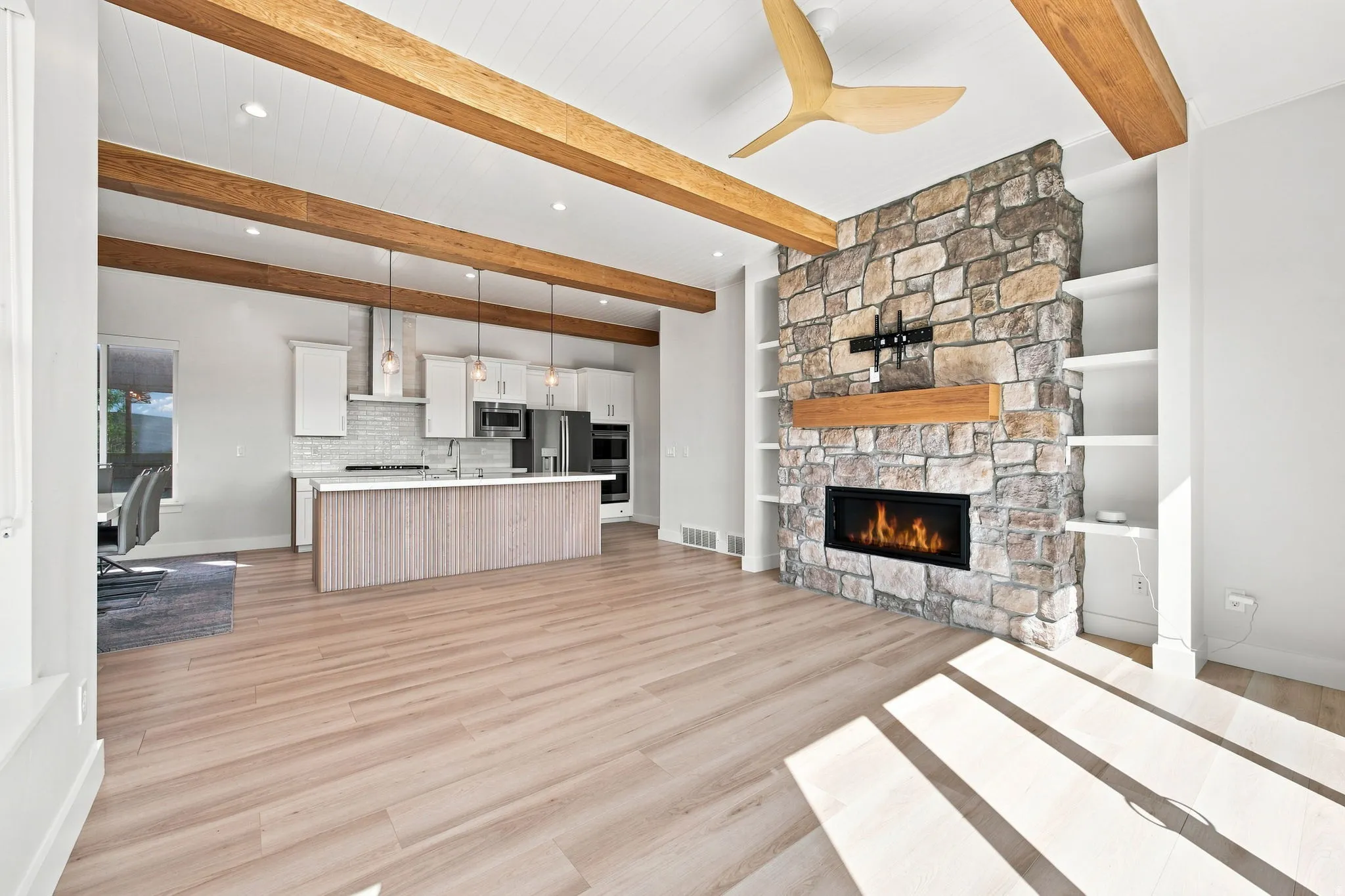 Stone fireplace with built-in shelving, wood-finish flooring, exposed ceiling beams, a modern ceiling fan, and a kitchen island with pendant lighting