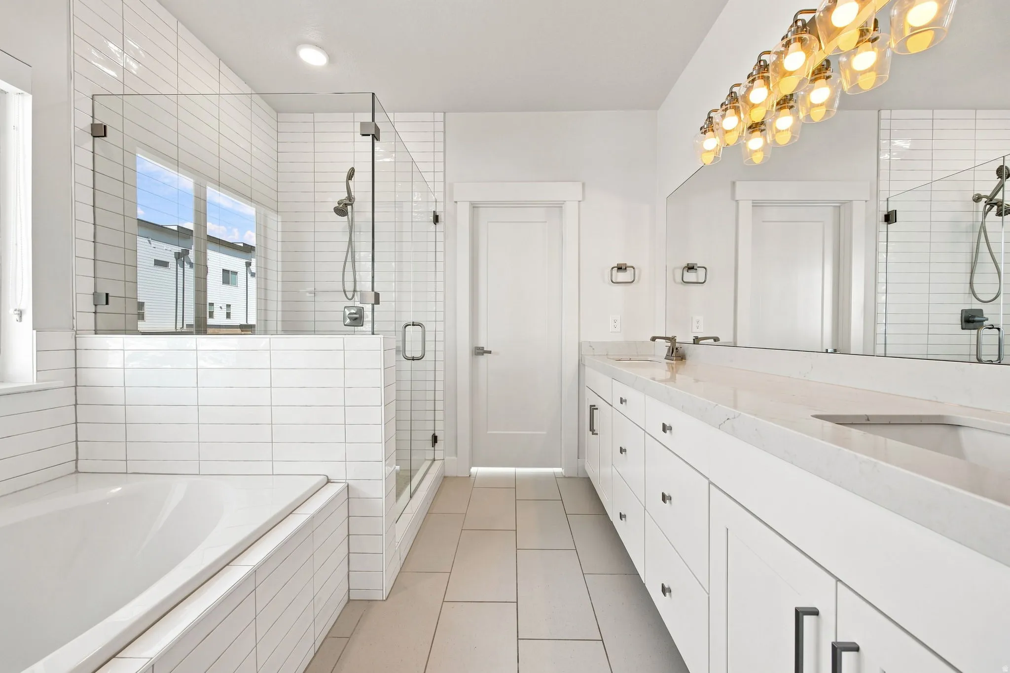 Glass-enclosed shower with subway tile surround, separate soaking tub, expansive vanity with dual sinks, and large mirror with decorative lighting