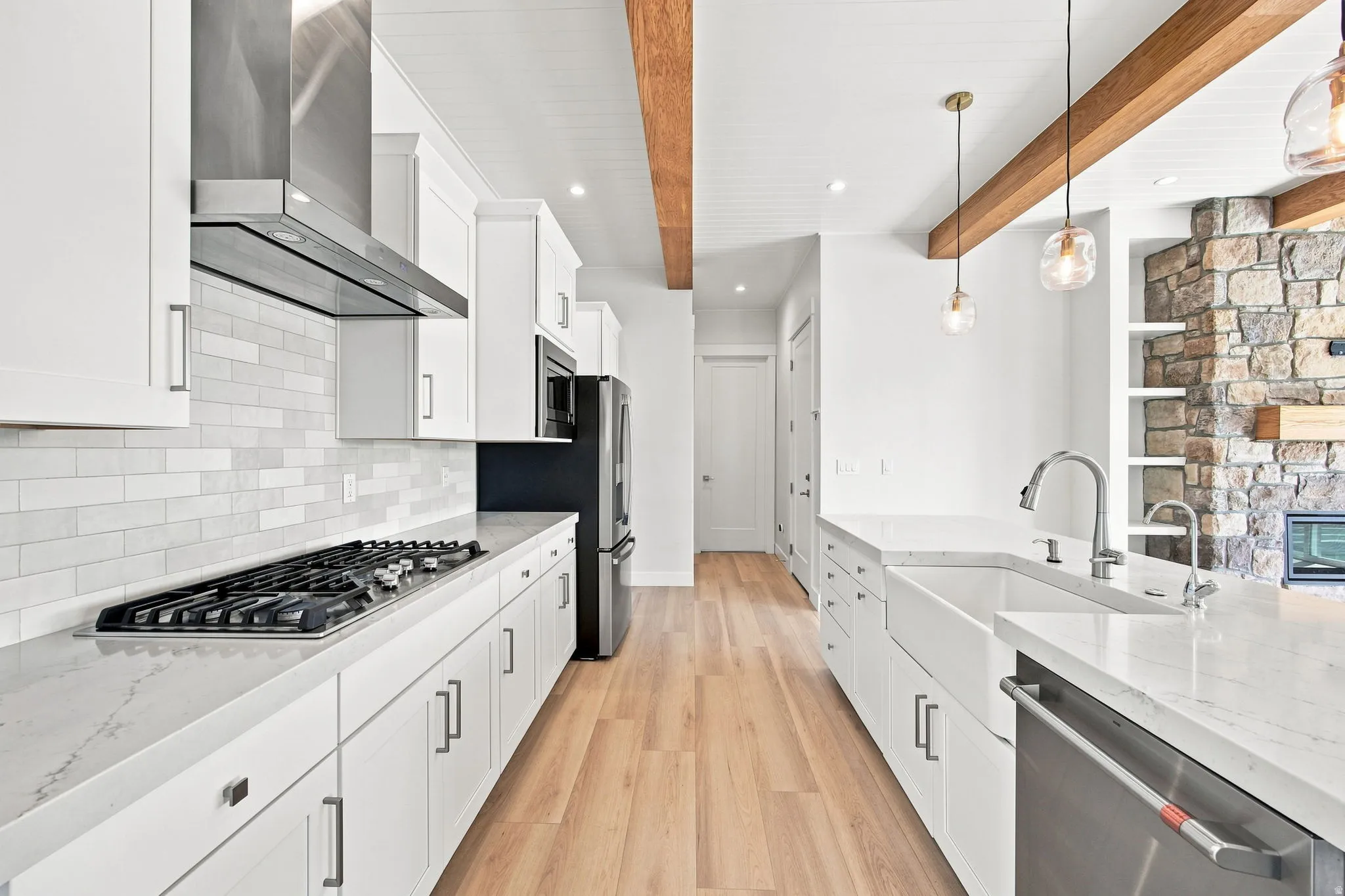 White shaker cabinetry, light wood-finish flooring, stone accent wall, stainless steel appliances, and exposed wooden beams