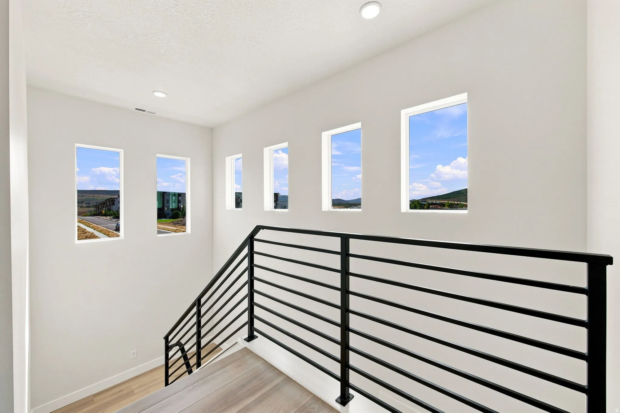 Modern staircase with black metal horizontal railings, light wood-finish flooring, and multiple rectangular windows