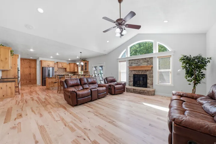 Vaulted ceiling with a ceiling fan, stone fireplace with a wood mantel, arched window, wood-finish flooring, and a kitchen with wood cabinetry