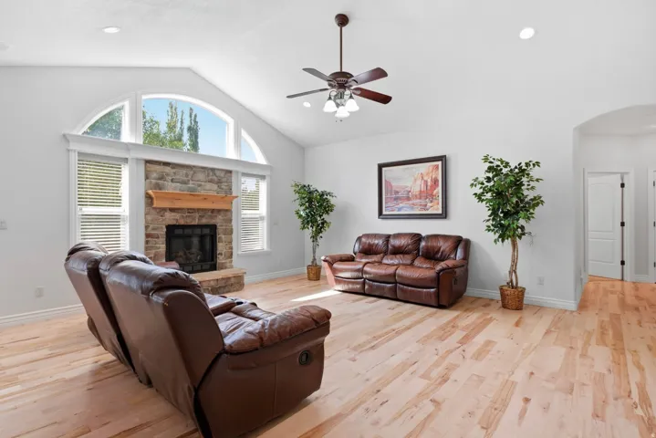 Vaulted ceiling with a ceiling fan, stone-faced fireplace with wood mantel, arched window, and hardwood-finish flooring