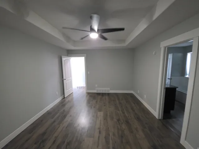 Tray ceiling with a ceiling fan, wood-finish flooring, light gray wall paint, white trim, and a built-in vanity in the adjacent room