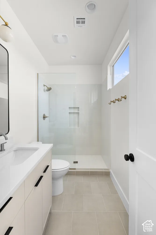 Walk-in shower with glass enclosure, single vanity with integrated sink, light-colored tile flooring, and a window for natural light