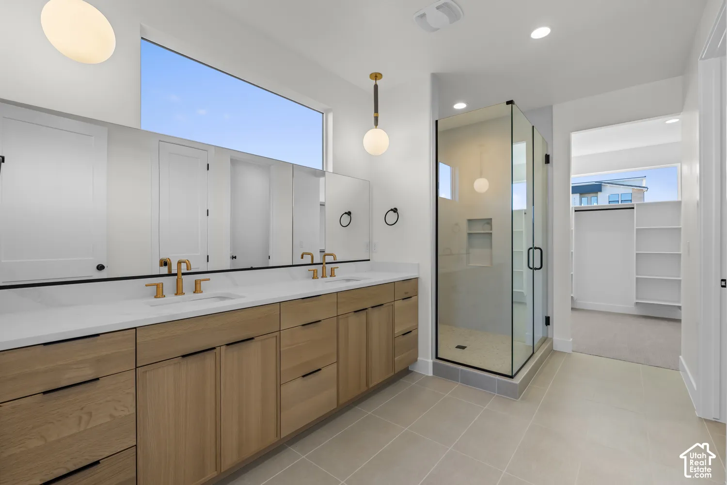 Dual vanity with brass fixtures and wood-finish cabinetry, frameless glass shower enclosure, large transom window, and neutral tile flooring