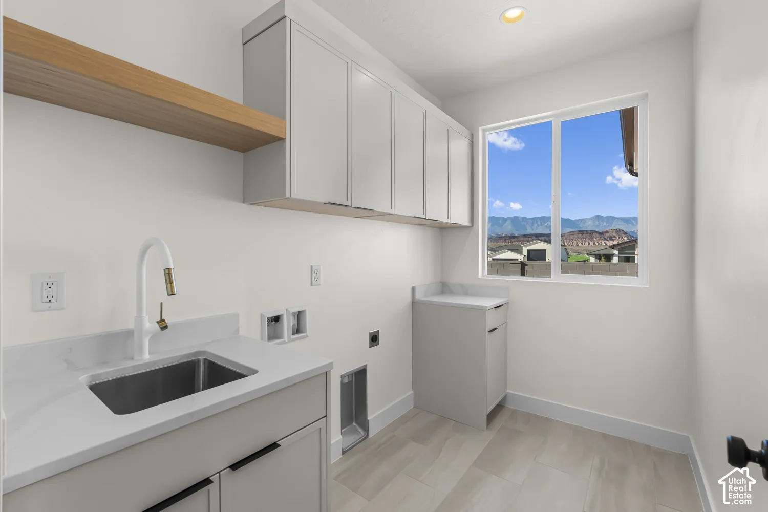 White cabinetry with black hardware, wood-finish shelving, an undermount sink with a white and gold-tone faucet, and a large window with mountain scenery