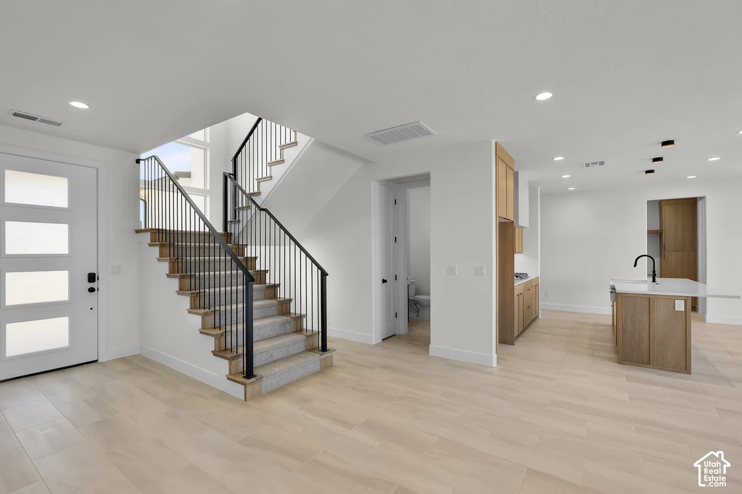 Light wood-finish flooring throughout, a modern staircase with black metal railings, a white front entry door with frosted glass panels, light wood-finish cabinetry, and a kitchen island with a white countertop and black faucet