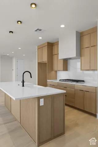 Kitchen featuring a central island with an undermount sink and matte black faucet, light wood-finish cabinetry, white countertops, and a stainless steel range hood