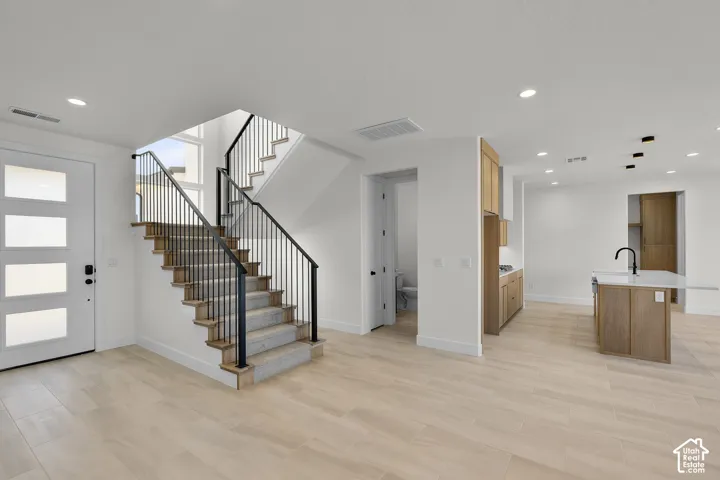 Light wood-finish flooring throughout, a modern staircase with black metal railings, a white front entry door with frosted glass panels, light wood-finish cabinetry, and a kitchen island with a white countertop and black faucet