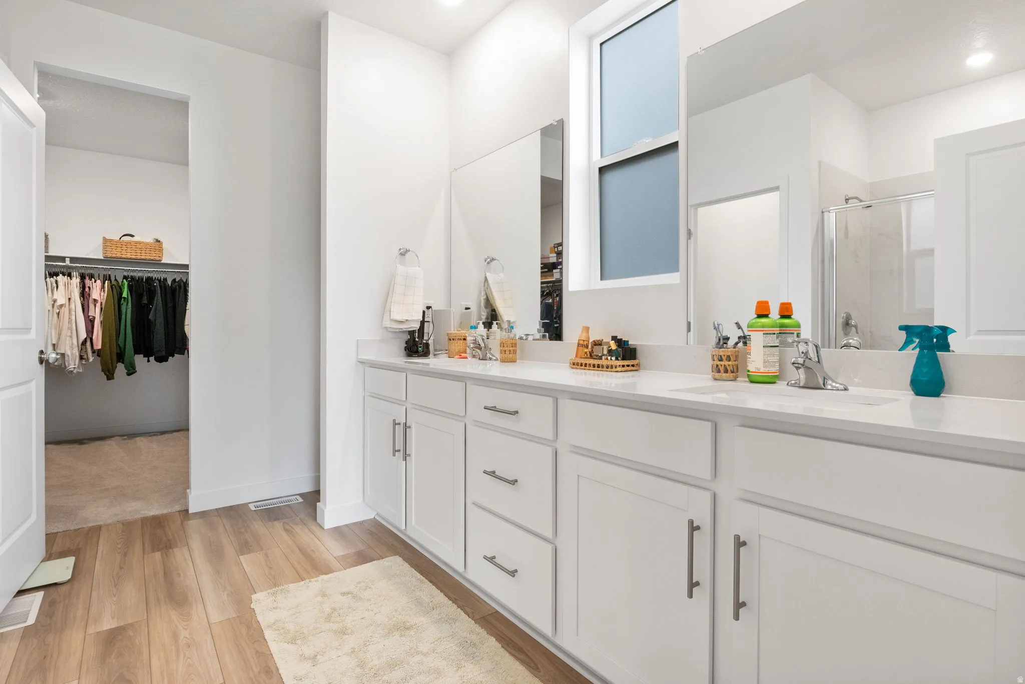 Large primary bathroom with dual vanit,  white cabinetry and integrated sinks, wood-finish flooring, and a walk-in closet.