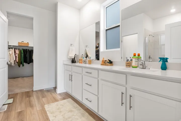 Large primary bathroom with dual vanit,  white cabinetry and integrated sinks, wood-finish flooring, and a walk-in closet.