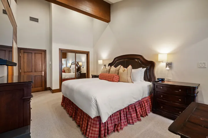 Vaulted ceiling with exposed wood beam, hardwood door, mirrored closet doors, built-in bedside sconces, and neutral carpet