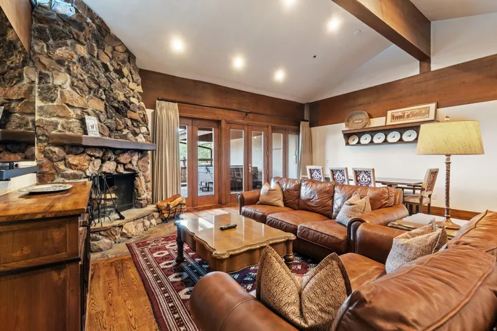 Vaulted ceiling with exposed wood beams, stone-clad fireplace with wood mantel, wood-finish flooring, and French doors