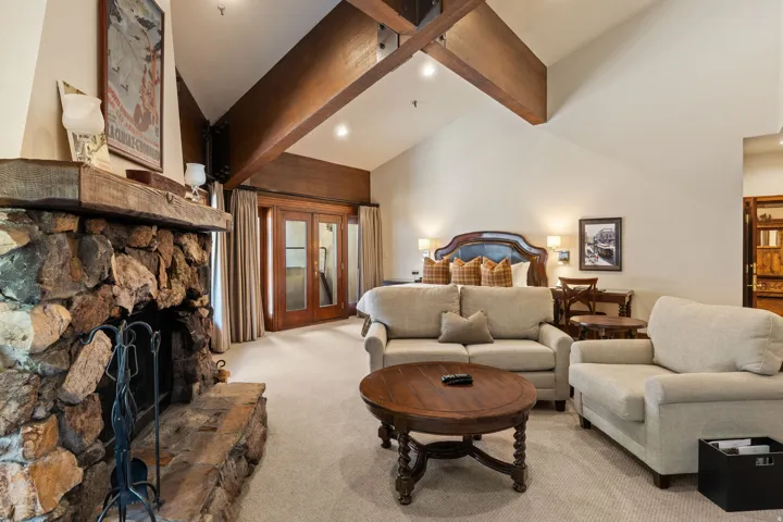 Spacious primary suite featuring exposed wooden beam ceiling, stone fireplace with fireplace tools, oval wood coffee table, and vaulted architecture with recessed lighting throughout