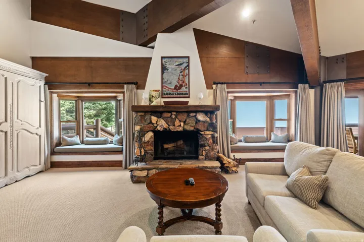 Vaulted ceiling with exposed wood beams, stone fireplace, built-in window seating, and carpeted flooring