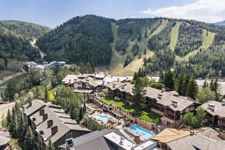 Expansive mountain resort community featuring multiple residential buildings with natural wood and stone exteriors, resort-style amenities including an outdoor heated pool, manicured landscaping with mature evergreen trees, and dramatic alpine backdrop wi