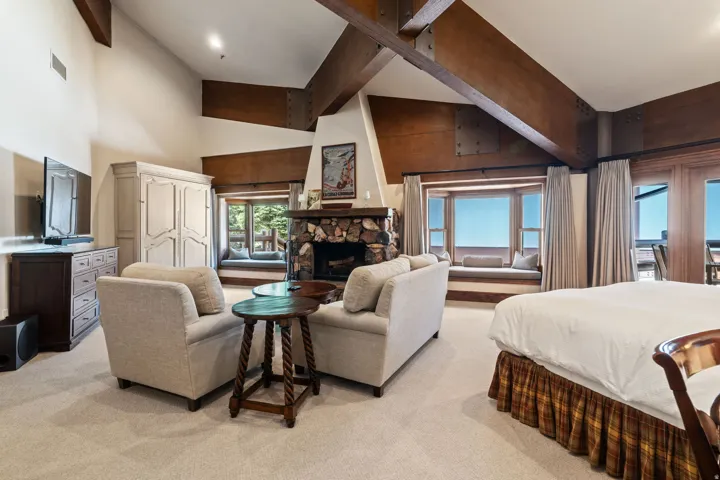 Vaulted ceiling with exposed wood beams, stone-clad fireplace, built-in window seating, and carpeted flooring