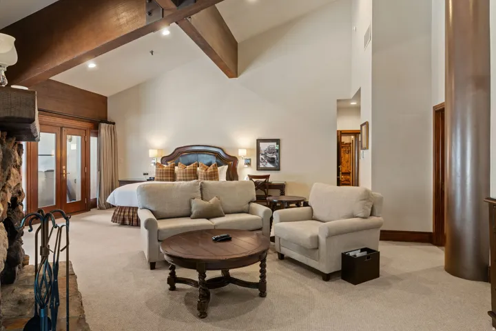 Vaulted ceilings with exposed wood beams, stone fireplace, light-toned carpeting, wood-finish French doors, and built-in wall sconces