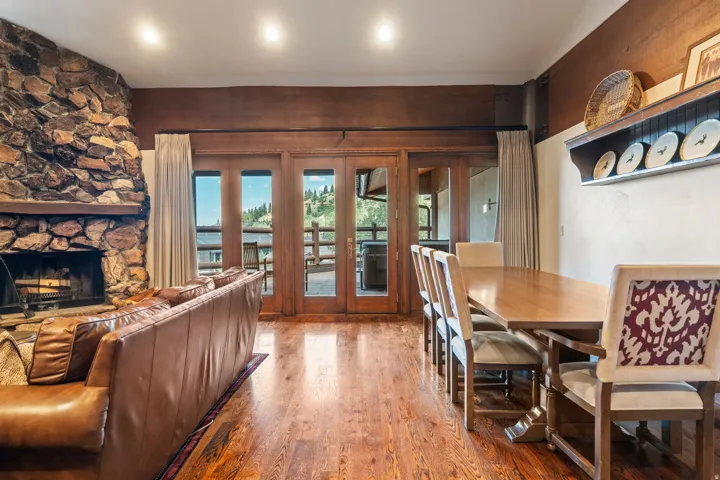 Stone-clad fireplace with wood-beam mantel, wood-finish flooring, and a bank of glass panel doors opening to an exterior deck