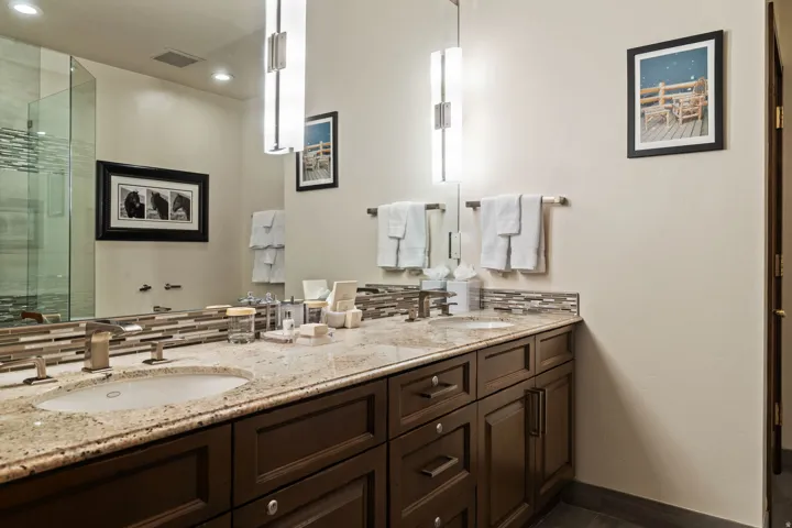 Dual undermount sinks, stone countertop, wood-finish cabinetry, brushed nickel fixtures, and a glass-enclosed shower
