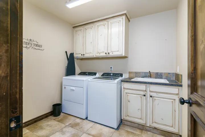 Laundry area featuring upper cabinetry, a utility sink with a stone tile backsplash, base cabinetry, and tiled flooring