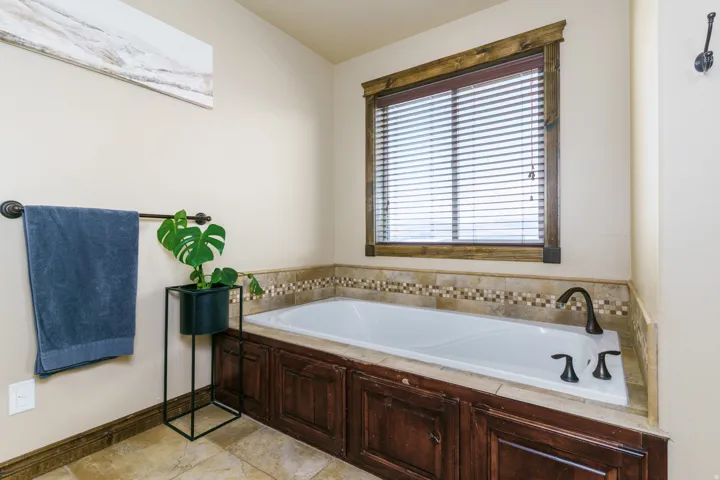 Built-in soaking tub with dark bronze fixtures, framed by a tiled surround featuring decorative mosaic accents, wood-finish paneling, and a wood-trimmed window