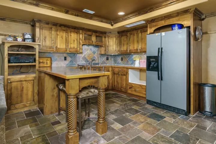 Custom wood cabinetry, stone tile flooring, built-in kitchen island, tile backsplash, and stainless steel refrigerator