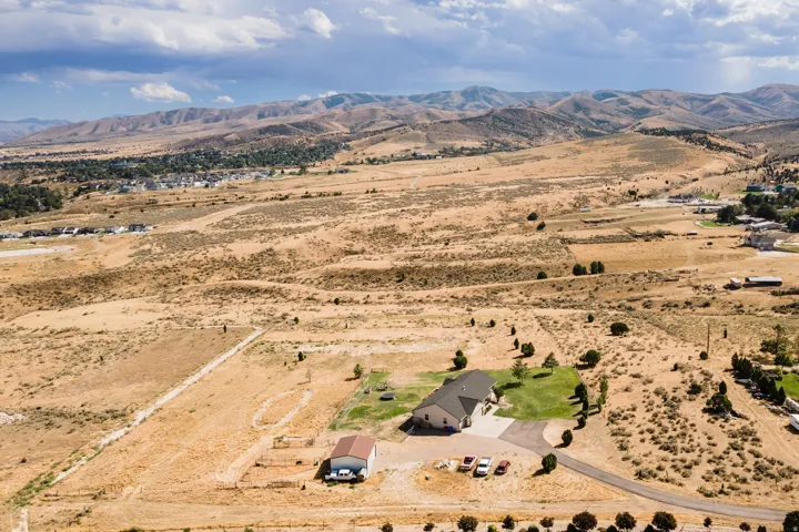 Expansive property featuring a single-story residence with a dark roof and light-colored siding, a detached outbuilding with a red roof, a circular driveway, a manicured lawn, and distant mountainous terrain