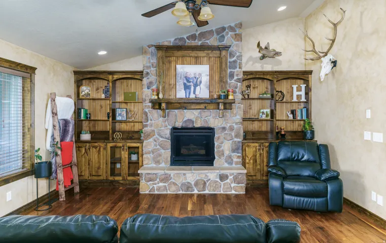 Vaulted ceiling with a ceiling fan, stone fireplace, built-in wooden shelving and cabinetry, and wood-finish flooring