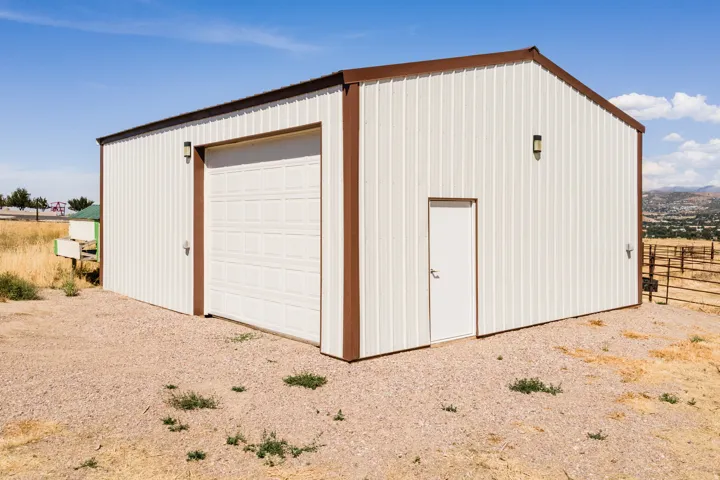 Metal-sided structure with brown trim, featuring a large panel garage door, a side entry door, and exterior wall lighting