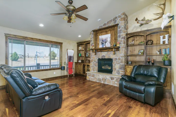 Hardwood-finish flooring, stone-clad fireplace with mantel, built-in shelving, ceiling fan with light fixture, and recessed lighting