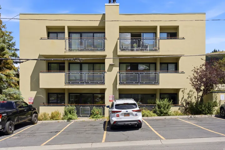 Multi-story building with a light-colored stucco exterior, featuring multiple balconies with metal and glass railings, and expansive dark-framed windows