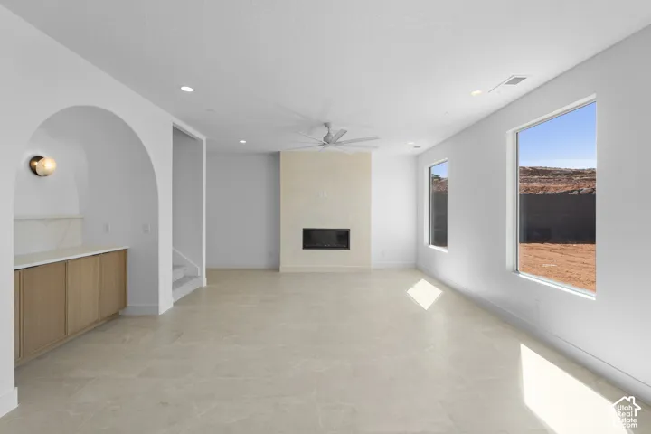 Spacious living area featuring light-toned tile flooring, a built-in fireplace, a ceiling fan, and large windows