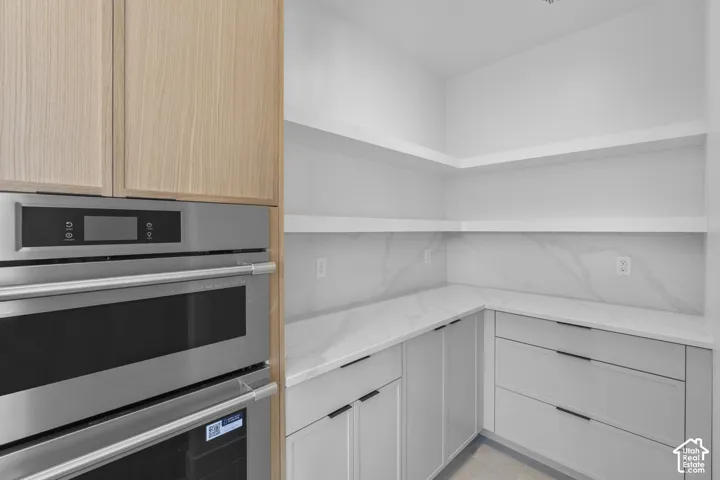 Built-in stainless steel double oven, light wood-finish cabinetry, white floating shelves, white countertops, and white lower cabinetry with matte black hardware