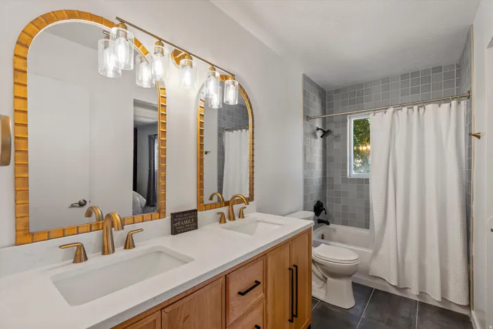 Dual arched mirrors with gold-tone frames, a vanity with a white countertop and two integrated sinks, gold-tone faucets, wood-finish cabinetry, and square tile wall surround in the shower