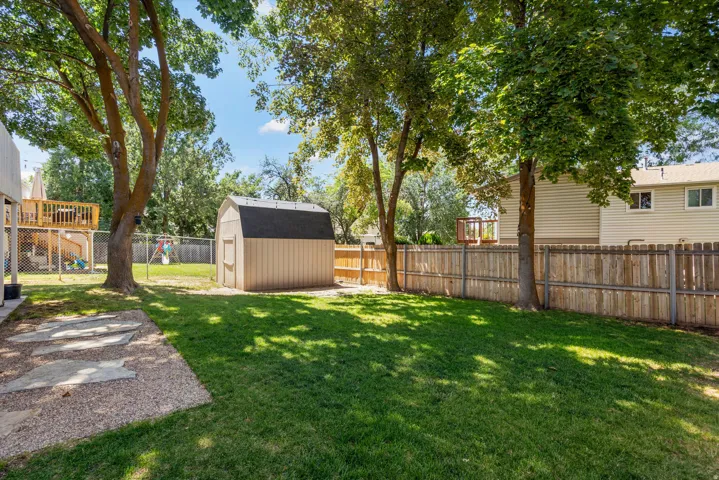 Expansive grassy yard featuring a wood privacy fence, mature trees, a storage shed, and a gravel patio with stepping stones