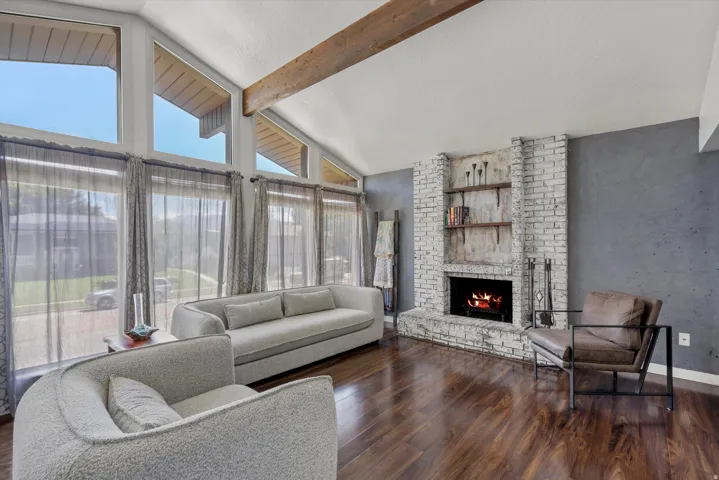 Vaulted ceiling with exposed wood beam, a brick-surround fireplace with built-in shelving, wood-finish flooring, and large windows