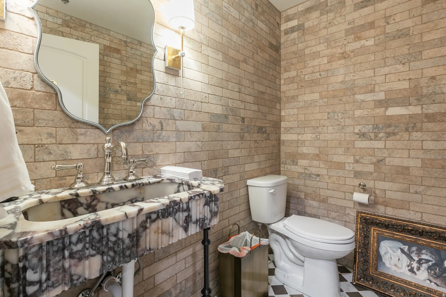 Scalloped vanity mirror, wall-mounted sconce lighting, marble-finish vanity with basin sink, checkerboard floor tiling, and brick-finish wall tiling