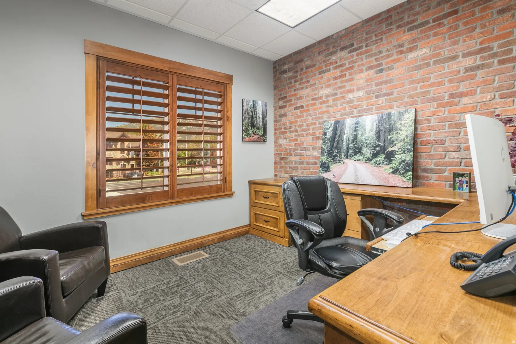 Exposed brick accent wall, wood-finish window shutters, wood-finish desk, wood-finish cabinetry, and patterned carpet