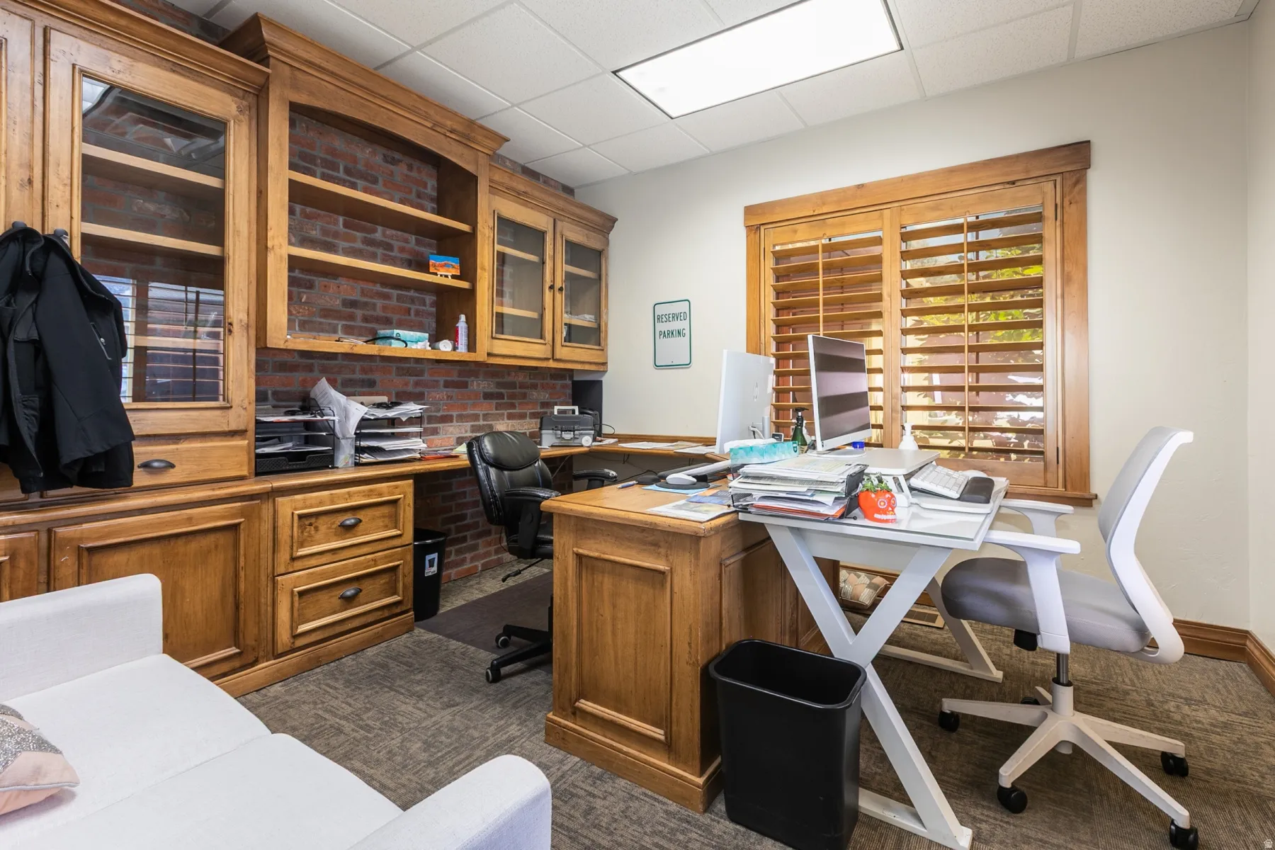 Built-in wood cabinetry with glass-front display cabinets, integrated wood desk, brick accent wall, wood-finish window shutters, and carpeted flooring
