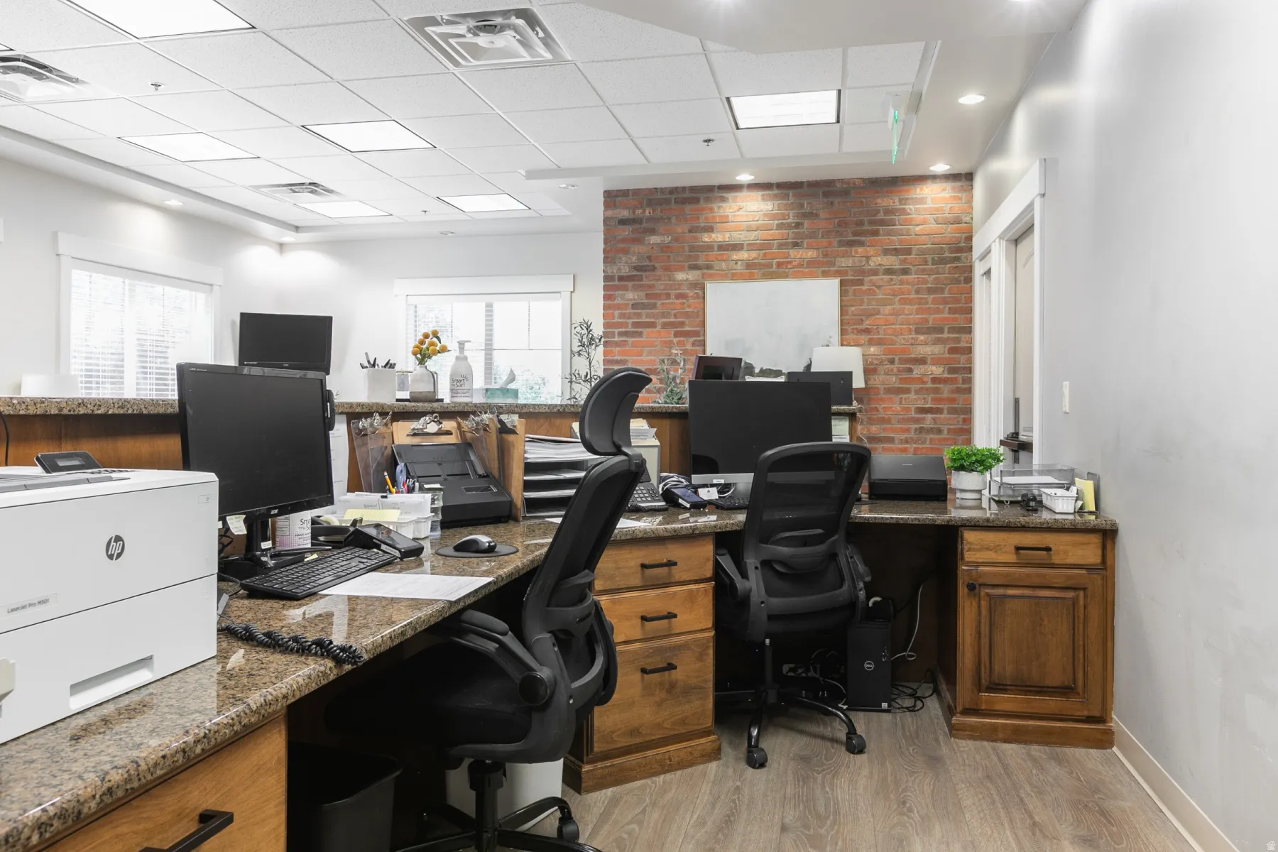 Professional office space featuring exposed brick accent wall with recessed lighting, wood-finish flooring throughout, multiple workstations with black ergonomic task seating, granite-style countertop surfaces, and ample built-in cabinetry with wood-finis