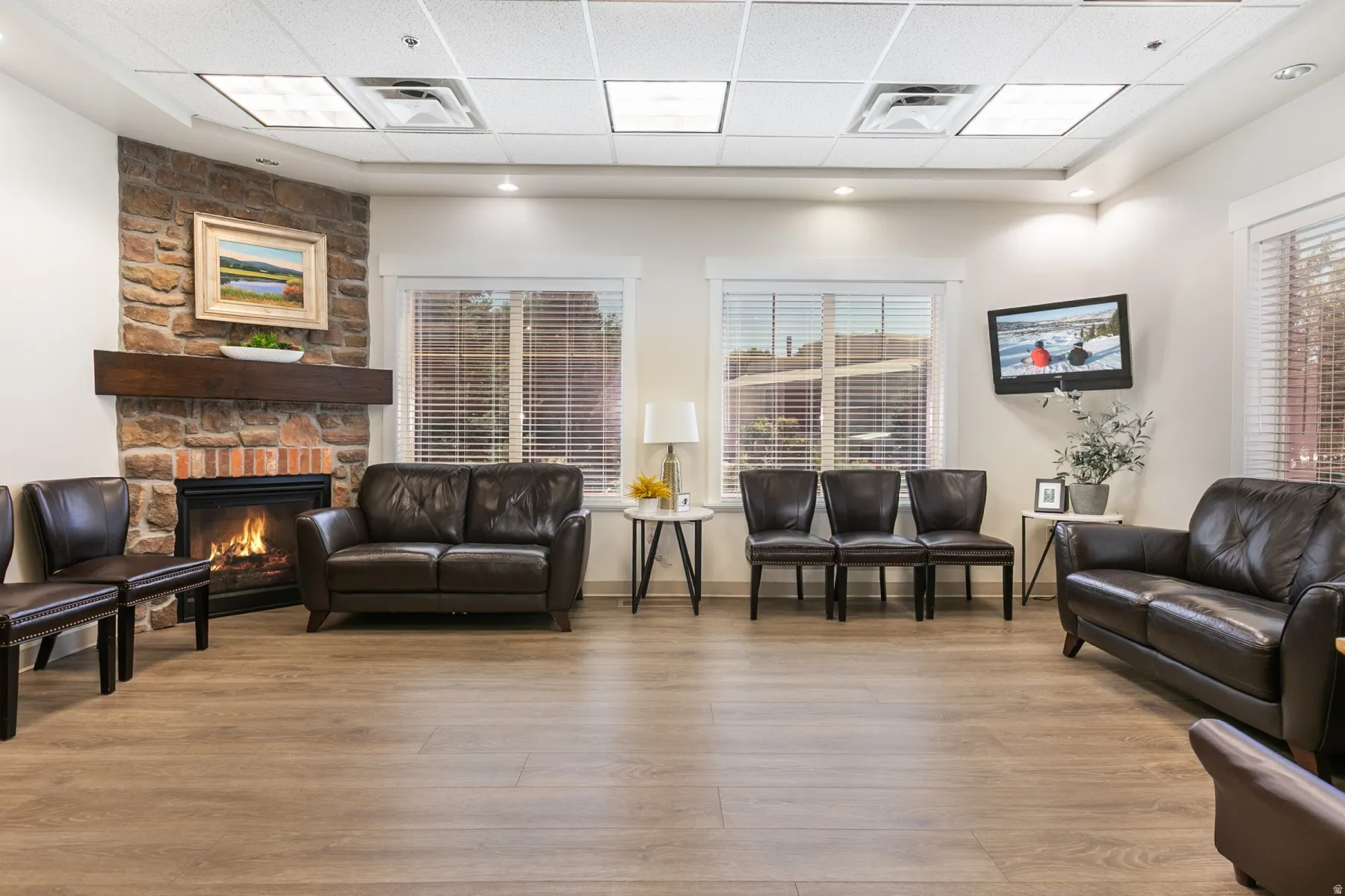 Stone-clad fireplace with a wood mantel, wood-finish flooring, recessed lighting, drop ceiling, and multiple windows