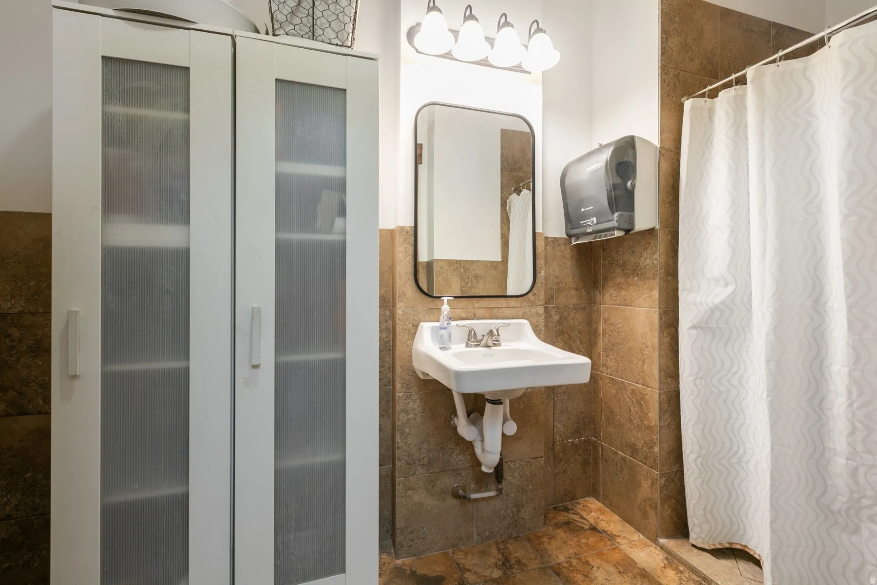 Bathroom featuring floor-to-ceiling tile, wall-mounted sink, shower with shower curtain, and a storage cabinet with frosted glass doors
