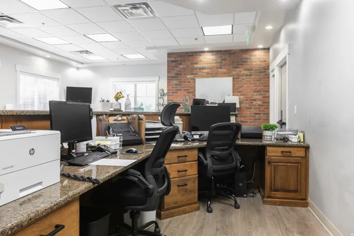 Professional office space featuring exposed brick accent wall with recessed lighting, wood-finish flooring throughout, multiple workstations with black ergonomic task seating, granite-style countertop surfaces, and ample built-in cabinetry with wood-finis