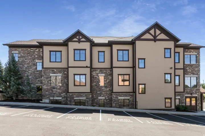 Multi-story building featuring stone and stucco exterior, prominent gabled architectural details, multiple windows, and an asphalt parking area with marked spaces
